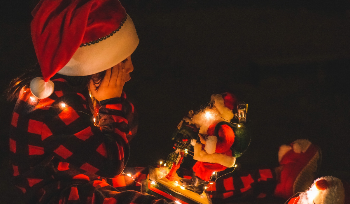 A young boy sitting in the dark with his legs out in front of him. He has a Santa Claus figure in his lap and holding his cheeks with his hands in disbelief at the lit up figurine. 